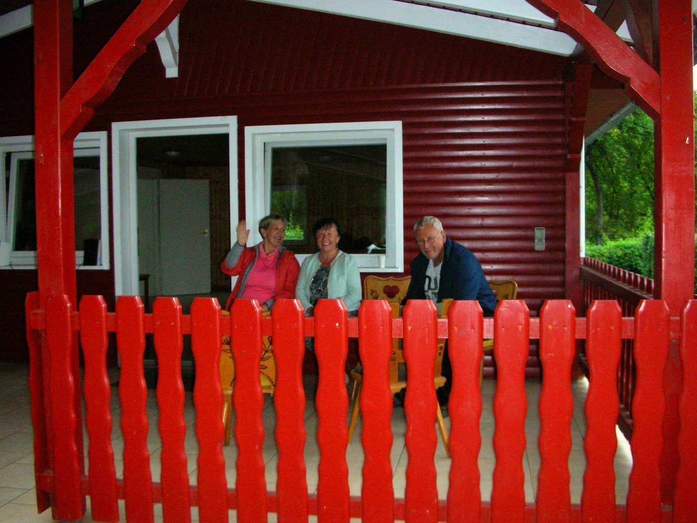Drei Gäste winken von der Veranda mit dem roten Holzgeländer